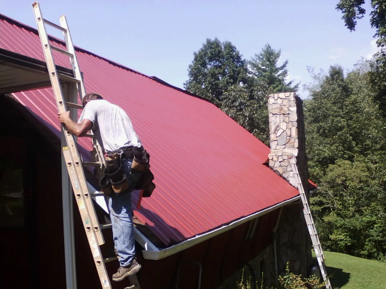 Roofer on a ladder beside a red metal roof and stone chimney — Soffit & Fascia Repair by Roofing Brothers in Bridgeville